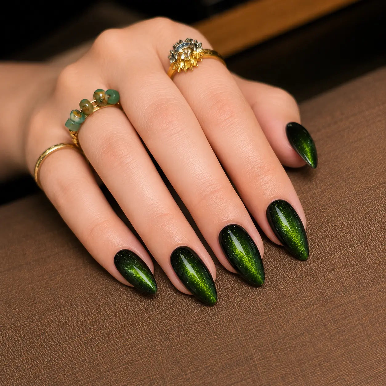 A close-up of a hand with long, almond-shaped nails painted in a metallic dark green ombre manicure, resting on a brown textured surface. The hand is adorned with gold rings.