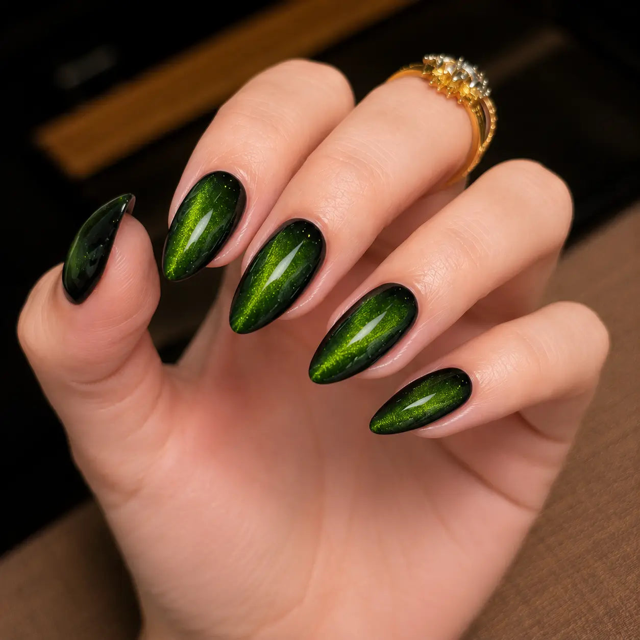 A close-up of a hand with long, almond-shaped nails painted in a metallic dark green and black cat-eye manicure, wearing a gold ring.
