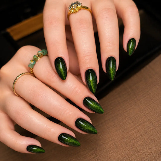 A close-up of two hands with long almond-shaped nails painted in a glossy dark green ombre manicure, styled with gold rings, against a brown textured background.